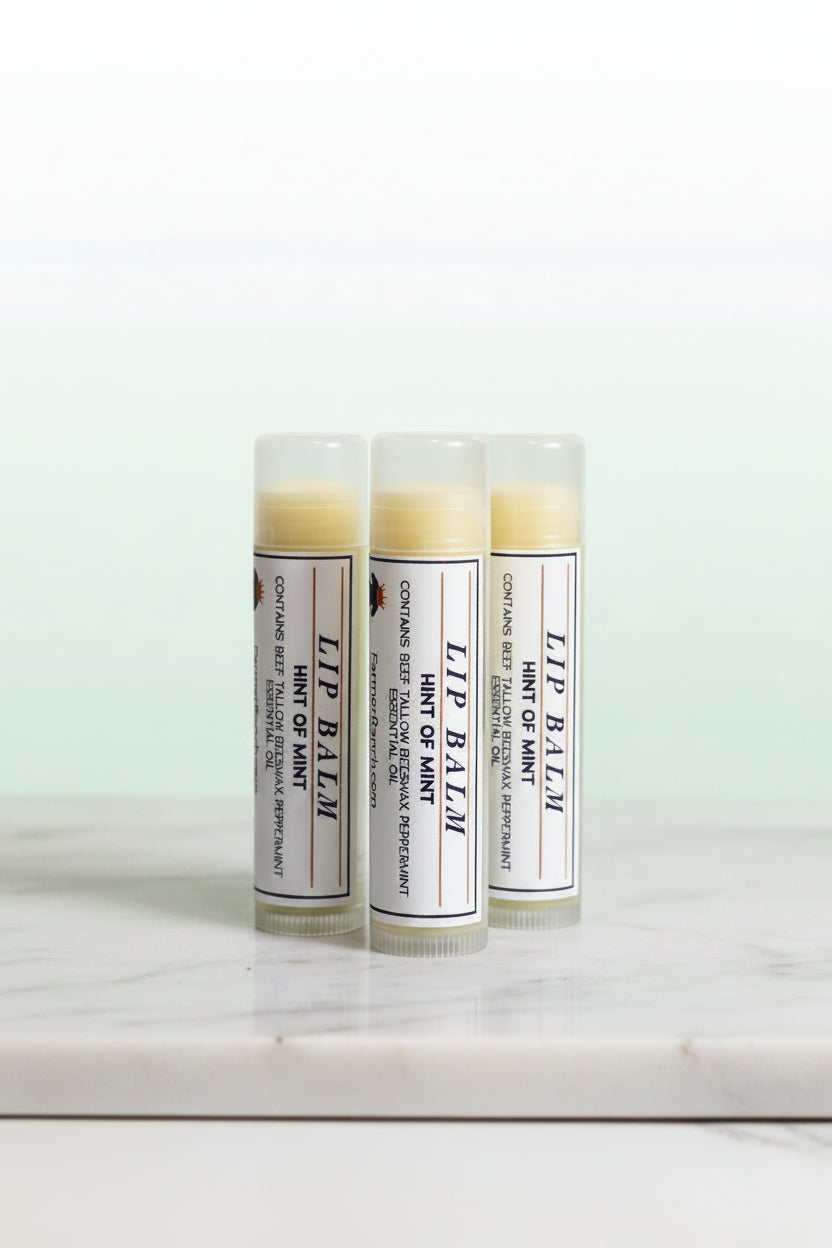 Three tubes of lip balm with white labels and balm visible at the top, standing on a white marble surface against a soft white background.