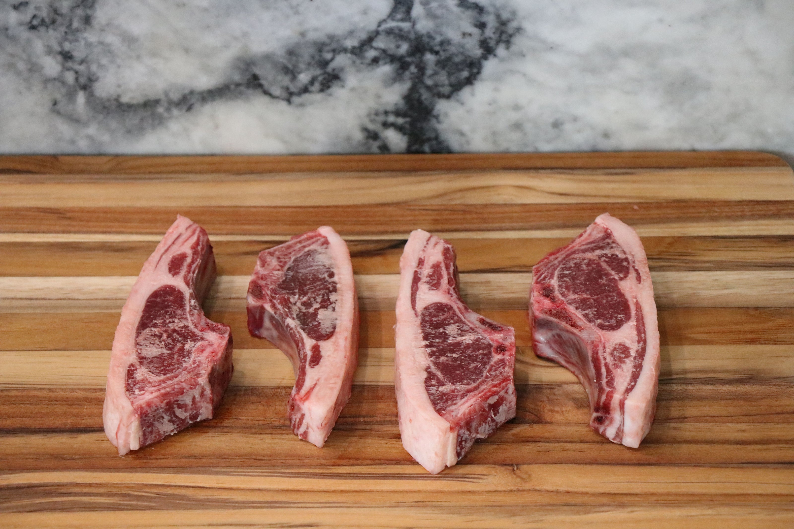 Four raw lamb chops with visible bone and marbling, arranged side by side on a wooden cutting board with a marble-patterned background.