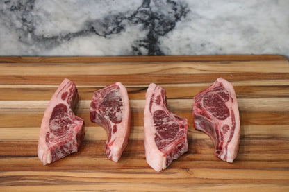 Four raw lamb chops with visible bone and marbling, arranged side by side on a wooden cutting board with a marble-patterned background.