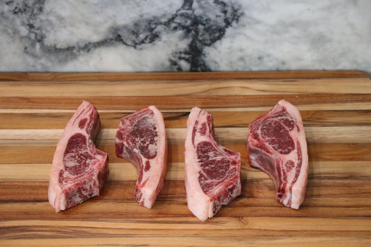 Four raw lamb chops with visible bone and marbling, arranged side by side on a wooden cutting board with a marble-patterned background.