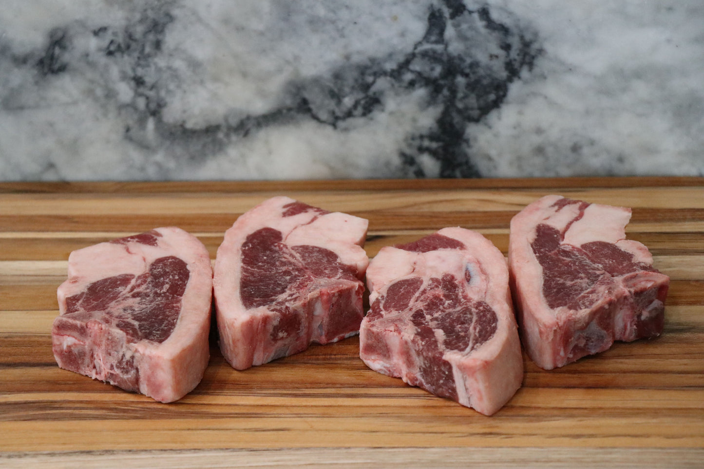 Four raw bone-in lamb chops with marbling, arranged on a wooden cutting board with a gray and white marble background.
