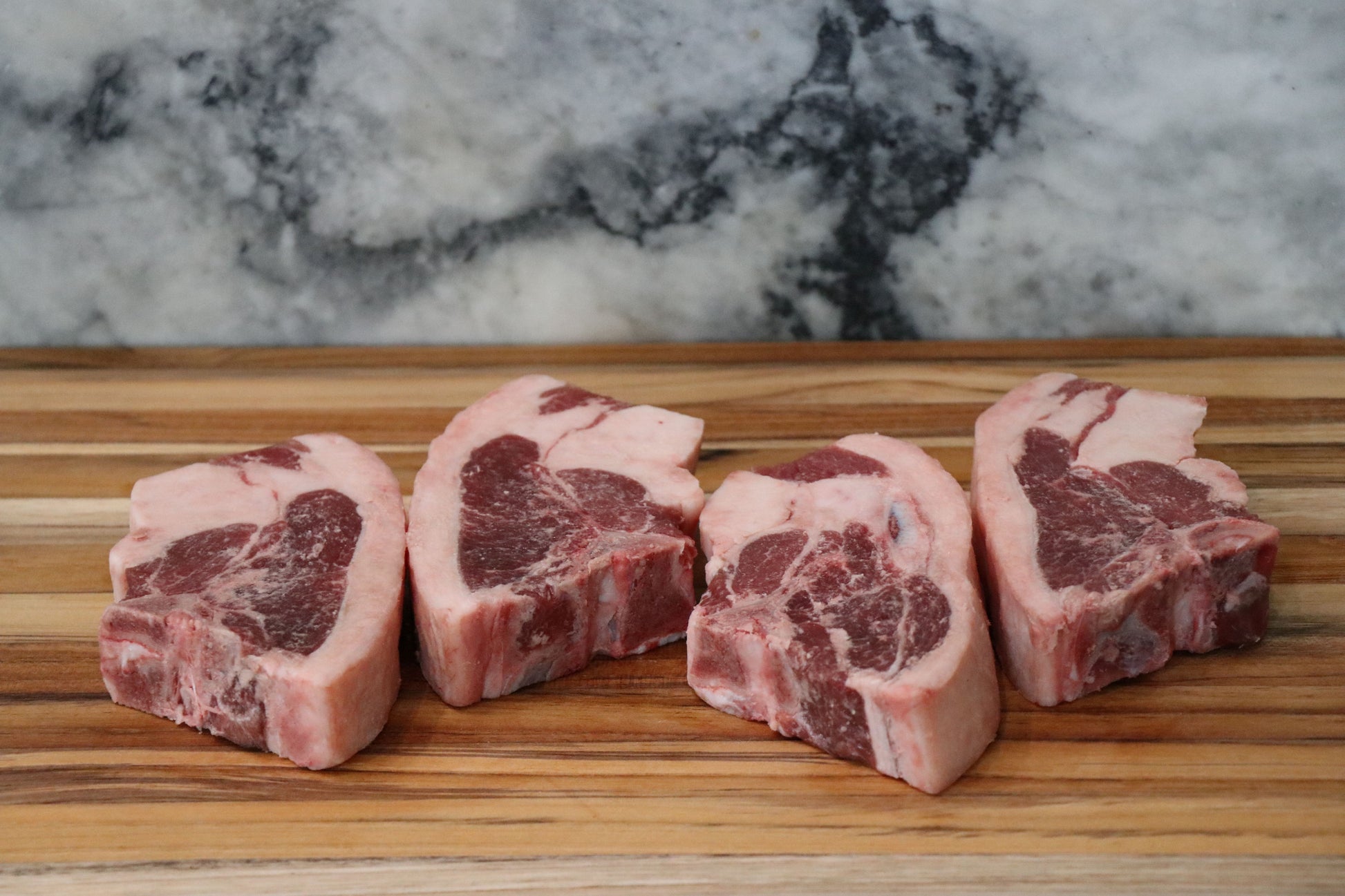 Four raw bone-in lamb chops with marbling, arranged on a wooden cutting board with a gray and white marble background.