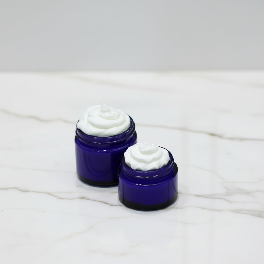 Two small open jars of white cream or balm in dark cobalt blue glass containers, placed on a white marble surface with a soft gray background.