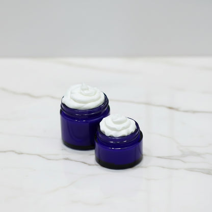 Two small open jars of white cream or balm in dark cobalt blue glass containers, placed on a white marble surface with a soft gray background.