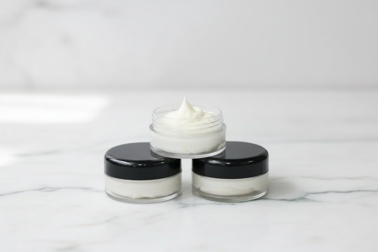 Three small cosmetic jars arranged in a pyramid on a white marble surface, with the top jar open to show a white cream or balm. The jars have black lids and clear plastic bodies.