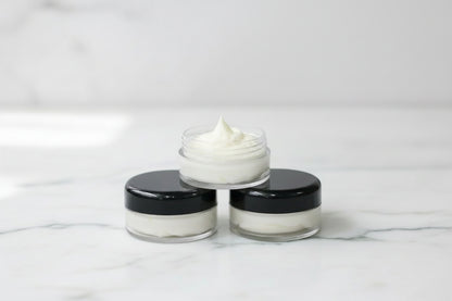 Three small cosmetic jars arranged in a pyramid on a white marble surface, with the top jar open to show a white cream or balm. The jars have black lids and clear plastic bodies.
