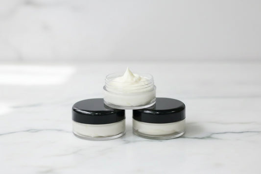Three small cosmetic jars arranged in a pyramid on a white marble surface, with the top jar open to show a white cream or balm. The jars have black lids and clear plastic bodies.